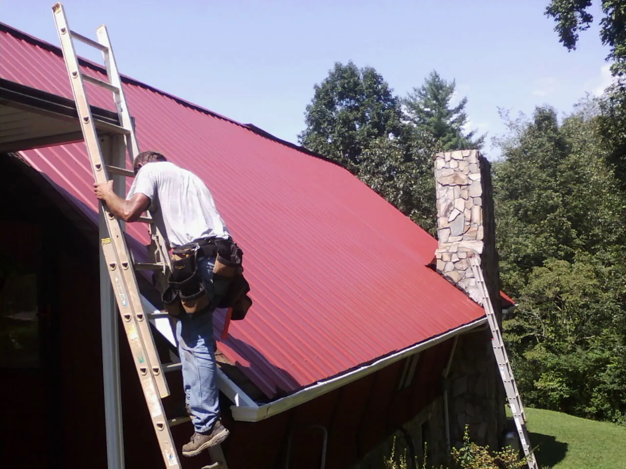 Roofer on a ladder beside a red metal roof and stone chimney — Soffit & Fascia Repair by Roofing Brothers in Westmoreland City